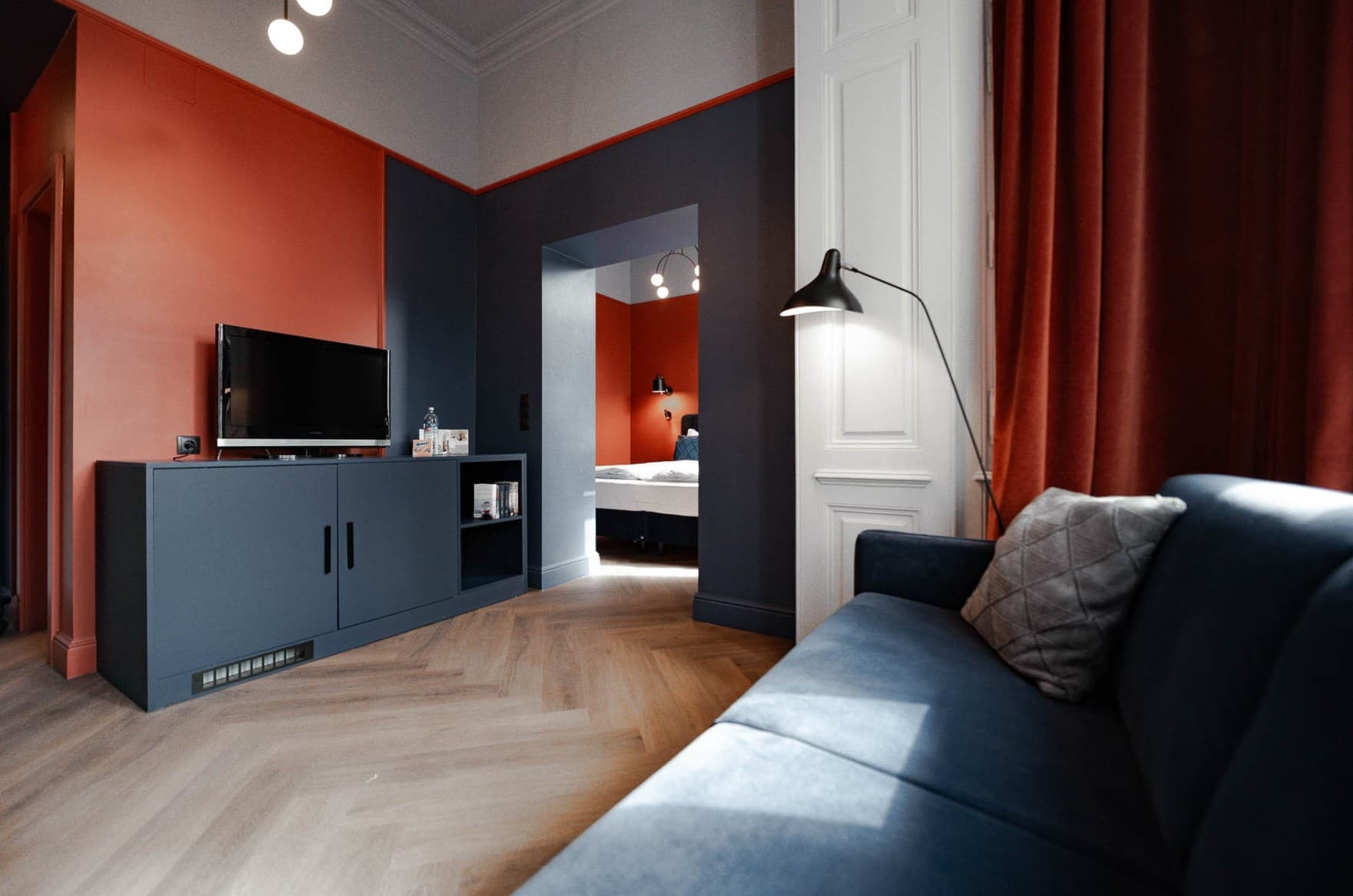 Seen is the Spacy category of the hideauts hotels Villa R, where you can see a couch in the living area in the front. In the back through a doorway, you can see the big double bed. It is styled in the art deco design, which has accents of dark blue and orange in the room.